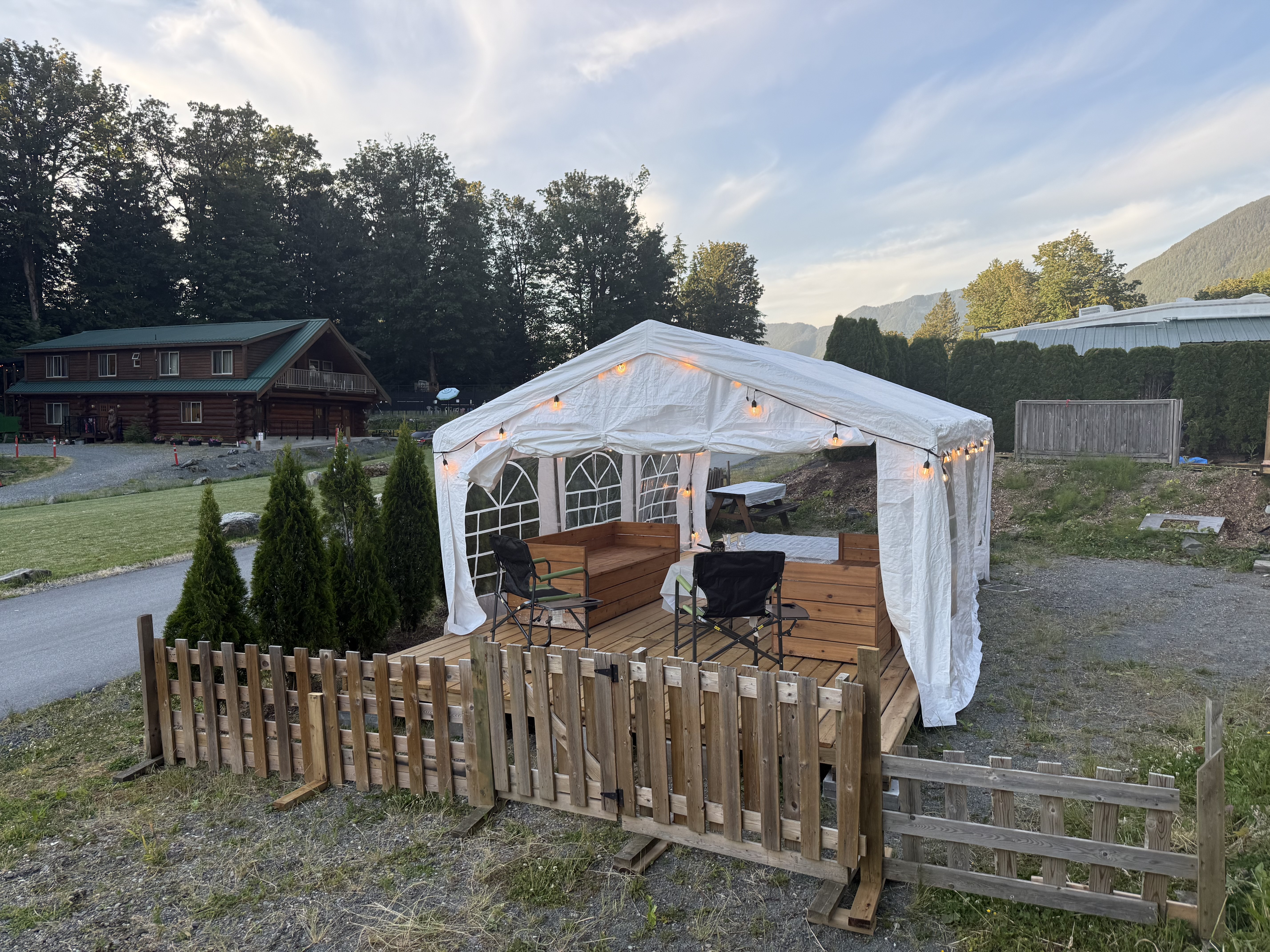 The Pavilion at dusk: a string-lit covered tent on a wood deck, the log lodge and Mt. Cheam behind